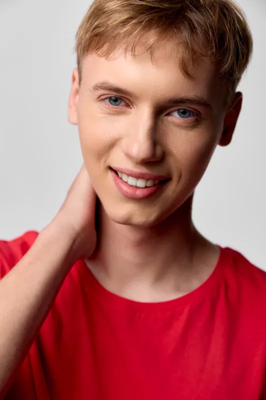 Smiling young male in a red shirt poses in a close up studio shot, hand resting at the neck, cheerful expression, bright lighting, casual headshot vibe