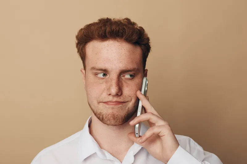 Young man with red hair and freckles wearing white shirt talks on smartphone with skeptical expression on beige background. Concept of communication, doubt, and casual lifestyle.