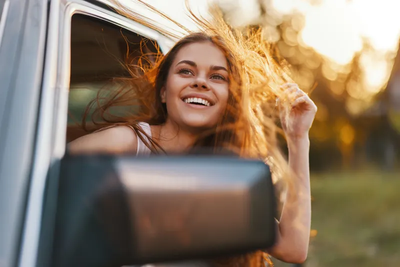 Happy young woman with long, flowing hair enjoying a sunny drive, smiling out of a car window amidst nature's vibrant colors and warm sunlight.
