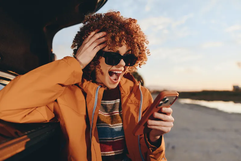 Woman with curly hair in orange jacket, holding a phone on a street, lifestyle outdoors, laughing near a car window at sunset, warm sunlight, candid portrait, young traveler, smiling selfie