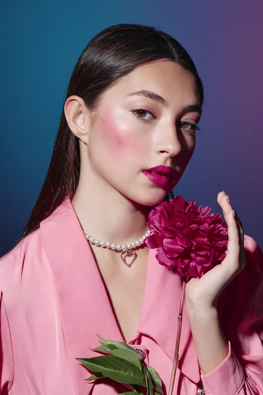 Young woman with glossy pink lipstick and shimmery cheeks holds a dark pink flower wearing a pink blazer with a pearl necklace. Studio beauty portrait on gradient background.