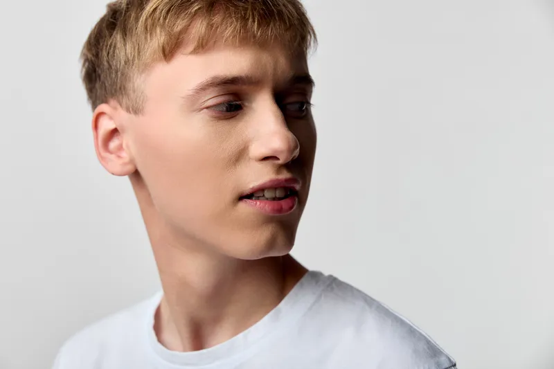 Young male with a side profile and calm expression, wearing a white t shirt, looking away. Minimal studio setup emphasizes natural mood and soft lighting.