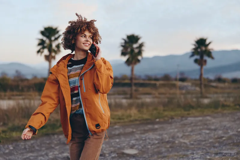 Lifestyle woman in orange jacket walks outdoors, smiling while talking on phone near palm trees, rural landscape and distant mountains at golden hour, casual communication concept, travel moment