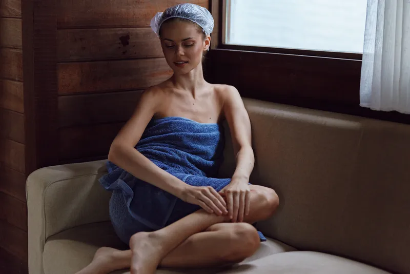 Relaxed woman wrapped in blue towel sitting on beige couch in cozy wooden room, wearing hair cap, enjoying calm moment by the window with soft natural light.