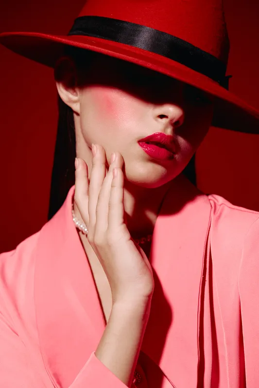 Woman with glossy red lips wears red hat that casts a shadow on her face. She poses with hand touching face, dressed in soft pink clothing, highlighting beauty, fashion, and creativity.