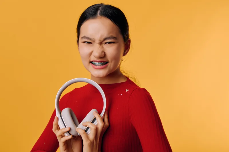 Young woman in red sweater holding headphones and smiling with braces on yellow background. Portrait, happiness, casual style and modern wireless technology for music and lifestyle.