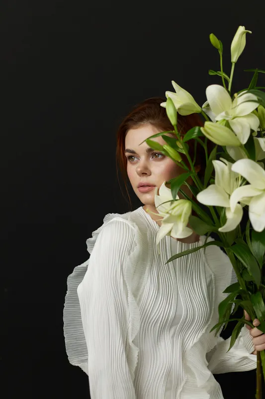 A serene young woman of Caucasian descent holding a bouquet of white lilies, showcasing elegance and tranquility against a dark background, perfect for wellness themes