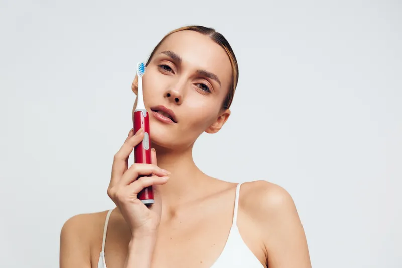 A young woman with clear skin holds a red electric toothbrush near her face against a plain white background. The concept focuses on personal hygiene and dental care.