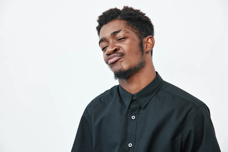 Young black man with dark skin wearing black shirt making confident face expression on white background. Portrait of African ethnicity male with short hair and casual style.
