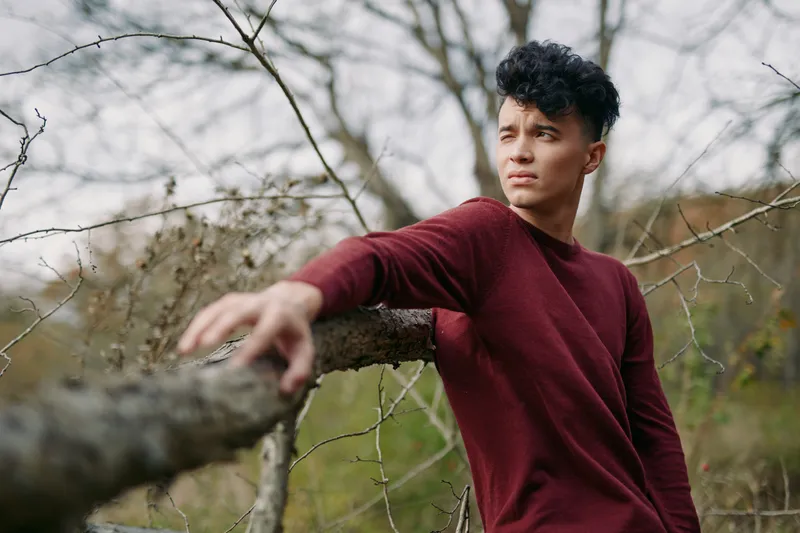 A candid portrait of a young man leaning on a fallen branch in a natural landscape, conveying authenticity and quiet strength through his thoughtful gaze and relaxed posture.