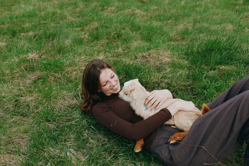 Casual lifestyle scene of a woman lying on grass, smiling, embracing a goat kid, outdoor calm, warm light, intimate moment, simple joy, peaceful connection with nature.