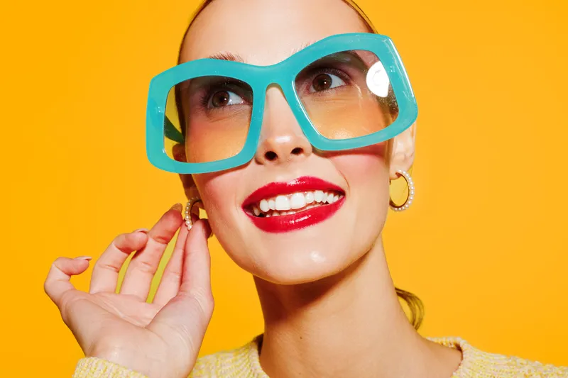 Smiling woman with large turquoise glasses and red lipstick poses against bright yellow background. Fashion portrait with vibrant colors and joyful expression. Concept of style and beauty.