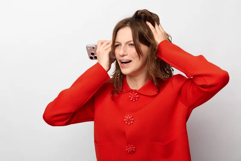 Happy woman in red sweater holds smartphone near ear, smiling and looking away. Isolated on white background. Concept for communication, technology, and positive emotions.
