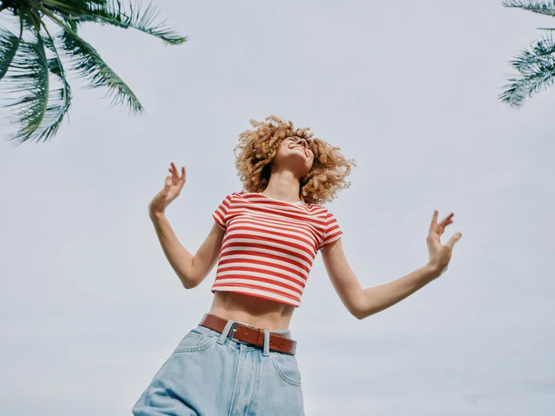 Young woman with curly hair wearing a striped crop top and denim shorts stands outdoors under palm trees, smiling with arms raised toward a clear sky.