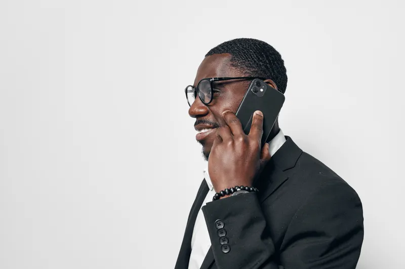 African American man with dark skin wearing glasses and black suit talking on smartphone in studio. Business professional, communication and technology concept with confident expression.