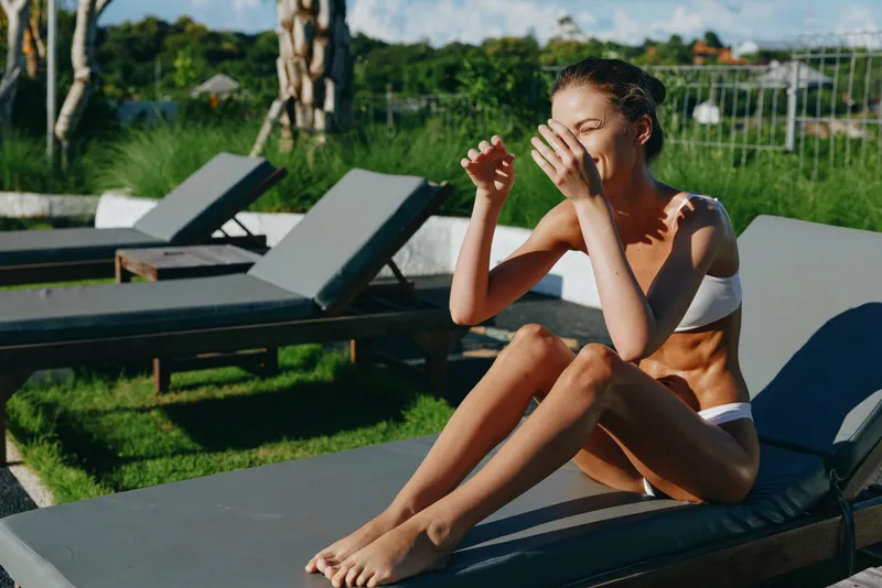 A woman in a swimsuit sits on a poolside lounge, wiping sweat from her face under bright sun. A sunlit outdoor relaxation moment by the pool in warm summer heat.