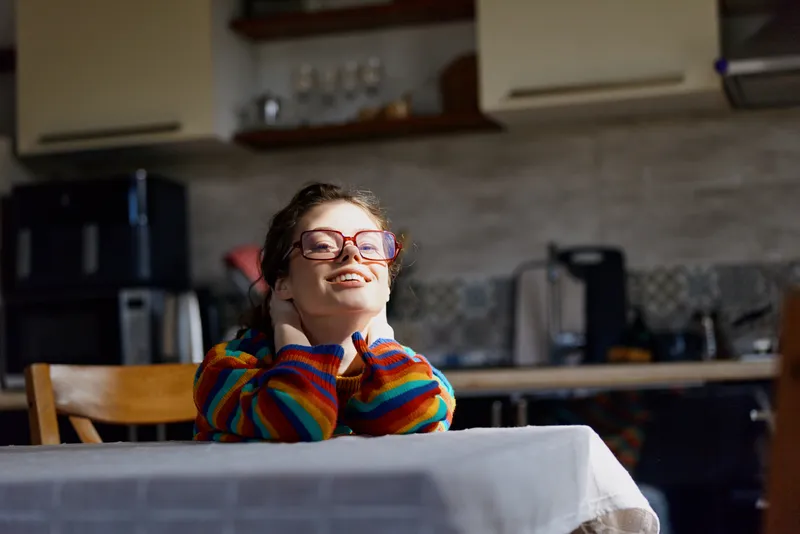 Cheerful woman indoors, relaxed pose at a dining table, colorful knit sweater, expressive glasses, soft natural light, cozy home vibe, candid lifestyle moment, bright kitchen backdrop