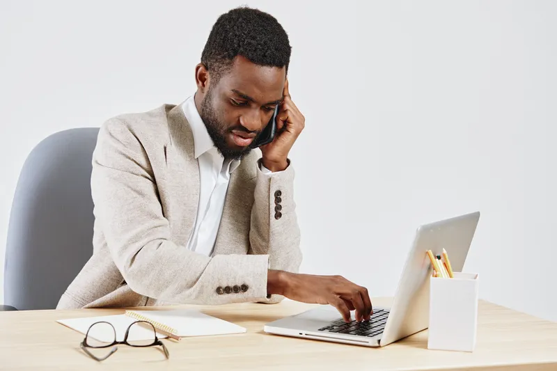 Professional young African American man in beige sweater at office desk using laptop and talking on cellphone, with glasses, notebook, pen holder, and minimal decor, on white background. Business and 