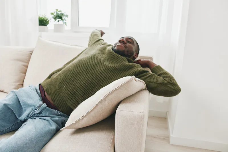 Relaxed African American man enjoying leisure time on a sofa, wearing a cozy green sweater, embodying comfort and serenity at home during the day
