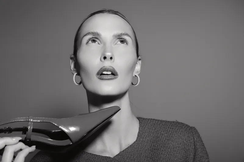 Close-up of a light skinned white woman holding a high heel shoe near her face. The studio portrait shows fashion concept, beauty, and elegance with soft lighting on grey background.