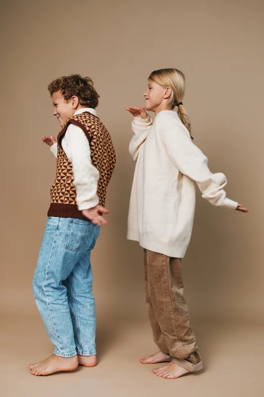 Two children dance playfully back to back in a studio setting, wearing cozy autumn outfits. Their smiles and carefree posture capture lighthearted energy and candid charm.