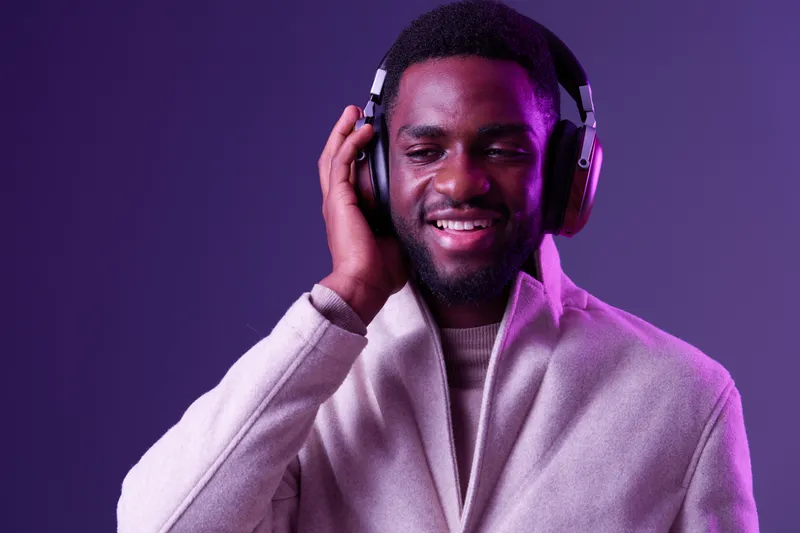 Smiling African man enjoying music with headphones, wearing a stylish coat, vibrant purple background, showcasing a modern and relaxed vibe