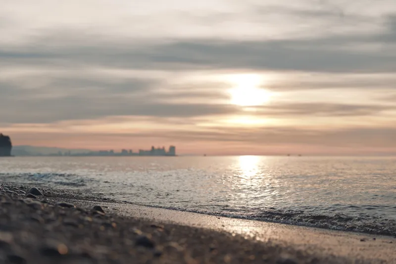 Beach at sunset with calm water, pebbled shore and distant city skyline on the horizon, warm light reflecting on waves and wet sand.