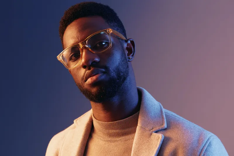 Stylish young Black man wearing glasses and a light sweater poses against a colorful gradient background, showcasing a confident expression and modern fashion sense