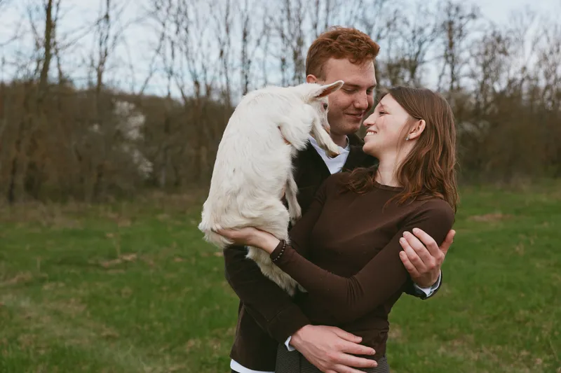 A couple embraces a white goat kid in a grassy field, smiling with warmth. Natural light highlights affection and countryside calm, a lifestyle moment focused on pet companionship and simple rural