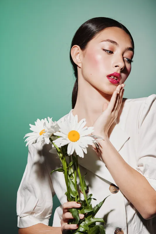 Young woman with sleek hair and glossy pink lips holds white daisies in one hand and touches her face softly on green background. Creative beauty and fashion concept with fresh look.