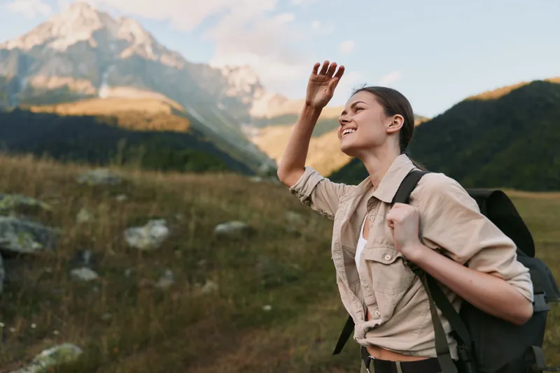 Young woman with brunette hair wearing a beige jacket and backpack enjoys a sunny mountain hike while smiling and shielding her eyes from the light.