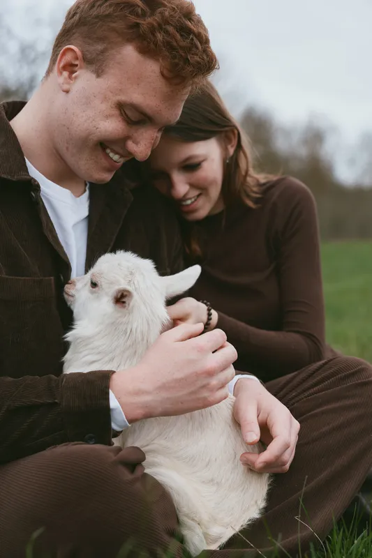 Young couple sitting on grass gently holding a white baby goat outdoors in a relaxed and affectionate moment during a casual lifestyle photoshoot.