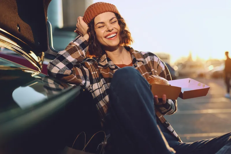 Woman smile car takeaway sunset casual joy candid portrait of a young female by a trunk holding a takeaway box in golden hour glow, authenticity and mindful living with emotional storytelling.