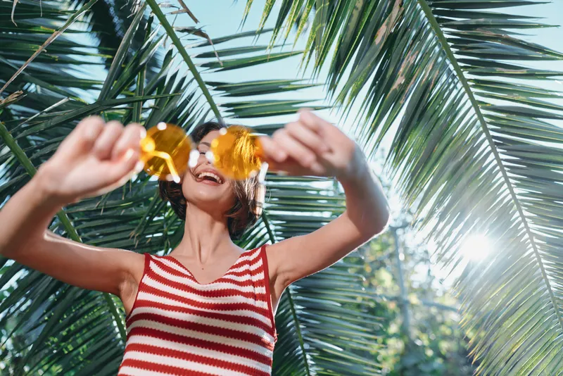 A cheerful woman wearing a red and white striped top smiles as she adjusts round sunglasses outdoors, surrounded by lush palm leaves and bright sunshine, creating a vibrant and carefree summer mood.