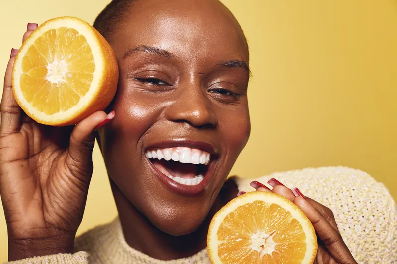 Smiling black woman with dark skin holds two orange halves near her face on yellow background. Concept of healthy lifestyle, natural beauty, vitamin C and skincare wellness.