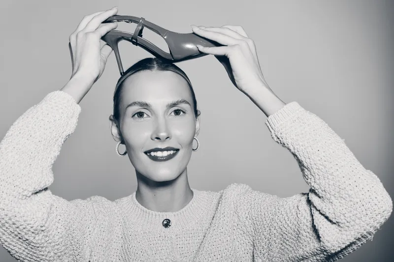 Smiling Caucasian woman with fair skin holds a high heel shoe above her head. Studio portrait in monochrome. Concept for fashion, beauty, and style accessories.