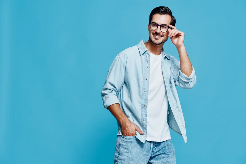 Smiling young man wearing casual outfit and glasses stands confidently against a bright blue backdrop, showcasing a relaxed and cheerful demeanor.