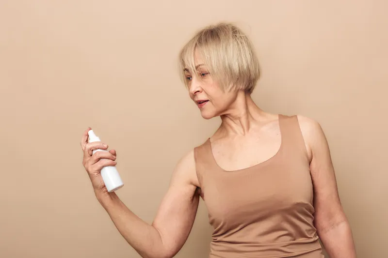 Mature woman with short blonde hair examines white spray bottle on beige background. Concept of skincare, self-care, aging beauty, and personal hygiene in natural light.