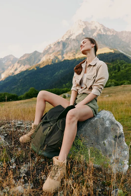 Casual outdoor woman rests on a rock in a mountain meadow, wearing a neutral shirt and backpack, enjoying expansive scenery and fresh air during a hiking day.