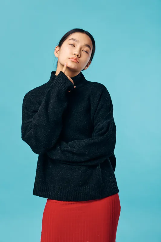 Young woman in black sweater and red skirt posing thoughtfully with hand on chin against blue background. Female model with calm expression and casual style for portrait photography.