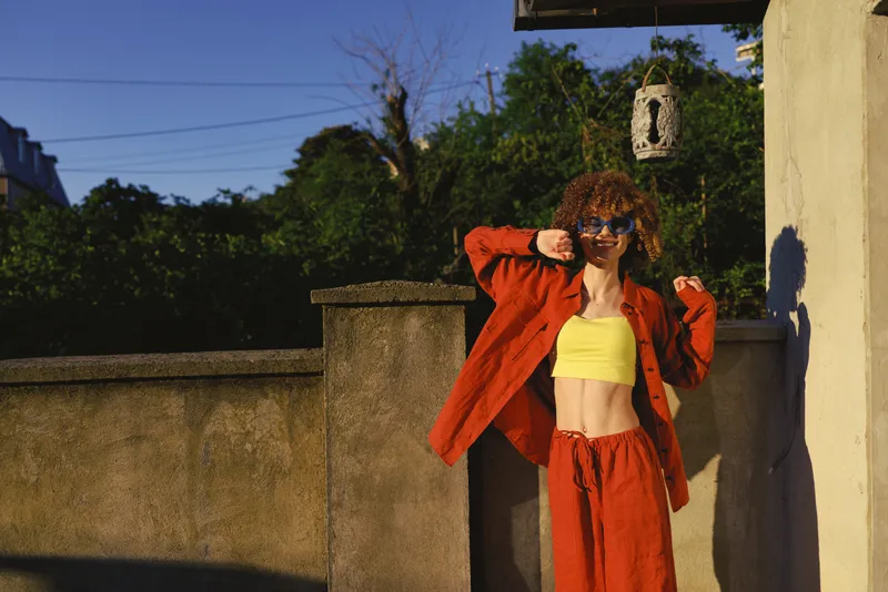Vibrant woman lifestyle scene outdoors with bright yellow crop top and orange jacket posing confidently in sunlight, casual street style portrait with warm tones and relaxed mood