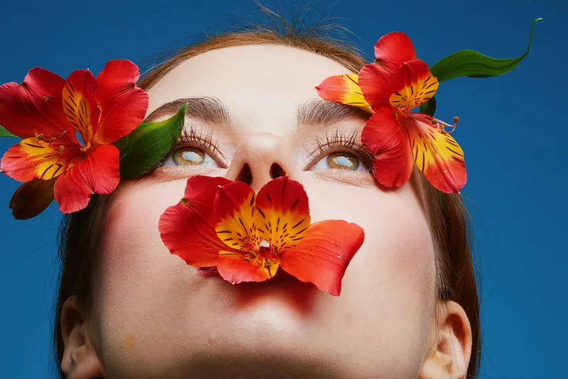 Portrait of a woman with vibrant flowers on her face, showcasing beauty, nature, and emotional expression against a clear blue background
