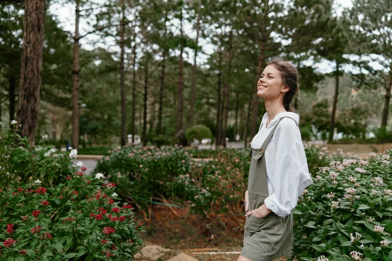 Young woman in casual outfit standing in flower garden, smiling joyfully, surrounded by greenery and vibrant blooms, representing happiness and nature