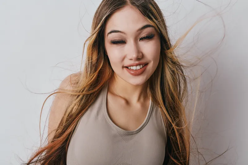 Young woman with long hair smiling with closed eyes against white background for beauty, fashion, portrait, happiness, and positive emotion in studio. Casual outfit and natural look.