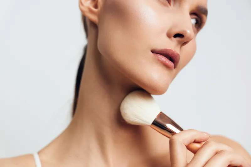 Young woman with smooth skin gently applies powder makeup on her neck using a large brush in a bright studio setting with a white background and soft lighting.