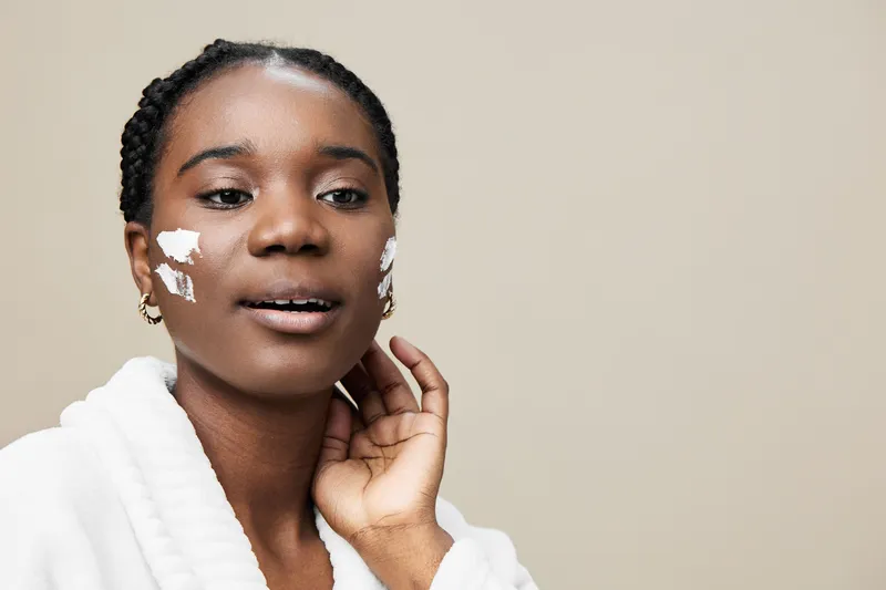 Young African woman in a white robe applying skincare cream on her face, showcasing a natural beauty routine against a neutral background
