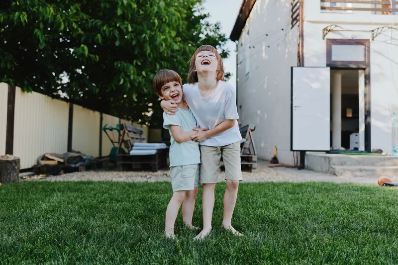 Two joyful boys smiling in a green yard, enjoying a sunny day. Their laughter reflects carefree childhood moments filled with happiness and playfulness.