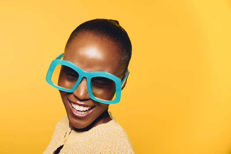 Happy African woman with dark skin wearing oversized blue sunglasses smiles joyfully against yellow background. Concept for fashion, summer, lifestyle, and positive emotions.