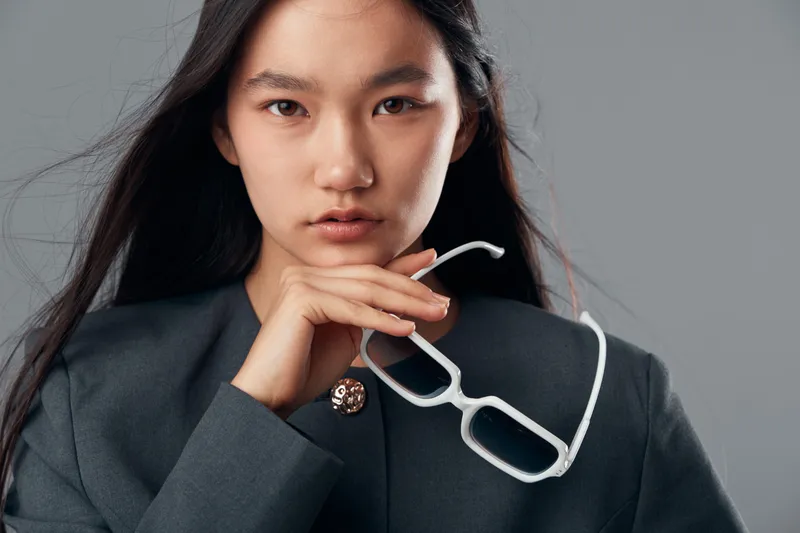 Young woman with long hair holding white glasses in hand wearing dark jacket looking at camera with serious face expression and neutral background in studio lighting.