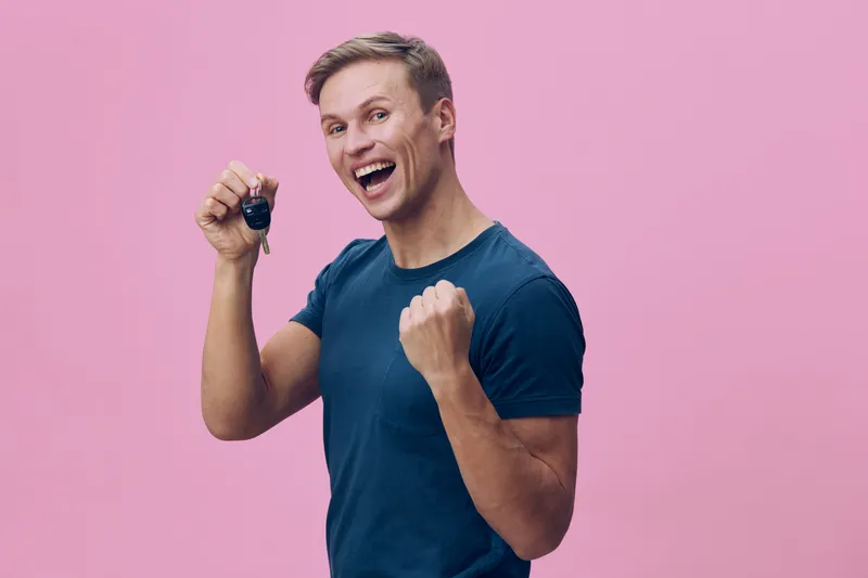 Cheerful young man holding car key fob smiling excited joyful celebrating success casual blue t-shirt pink background studio portrait. People lifestyle concept